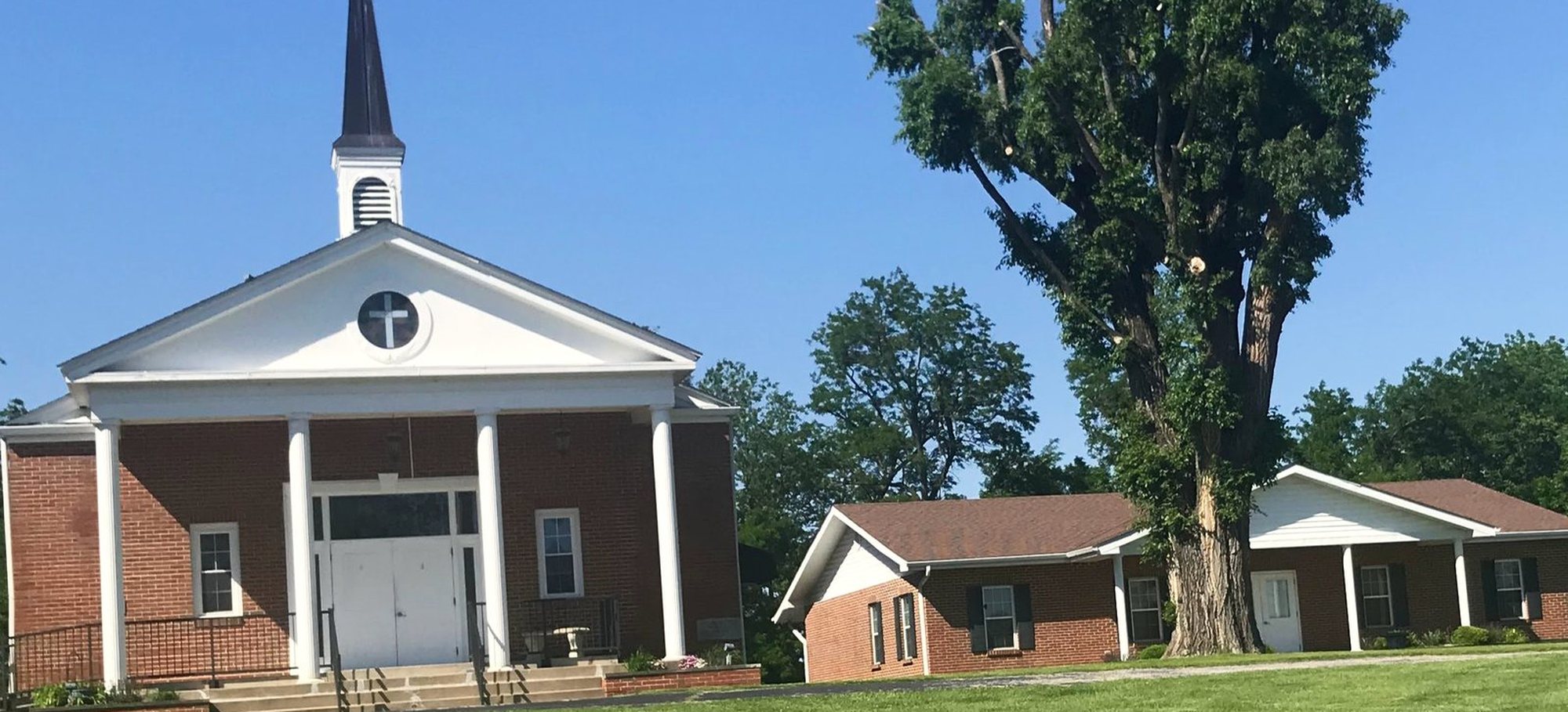 Temple Christian Fellowship on a clear summer morning: a red brick sanctuary with four white columns, a white portico and a dark steeple topped by a cross, with a low brick fellowship hall to the right beneath a large old tree, and a wide green lawn in front.