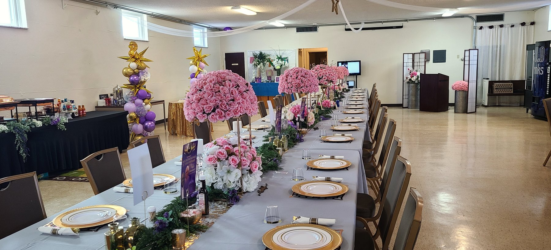 The fellowship hall laid out for a banquet: one long table dressed in white and gold with gold charger plates, tall arrangements of pink and white flowers down the centre, and chairs set for a full room.