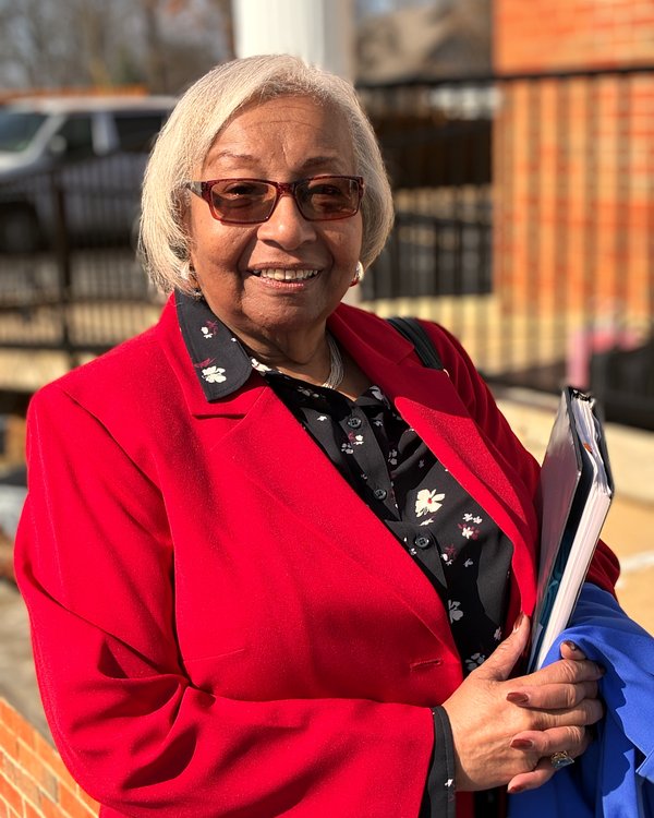 Elder Jeanette McNutt, smiling, in a red jacket, holding a folder outside the church.