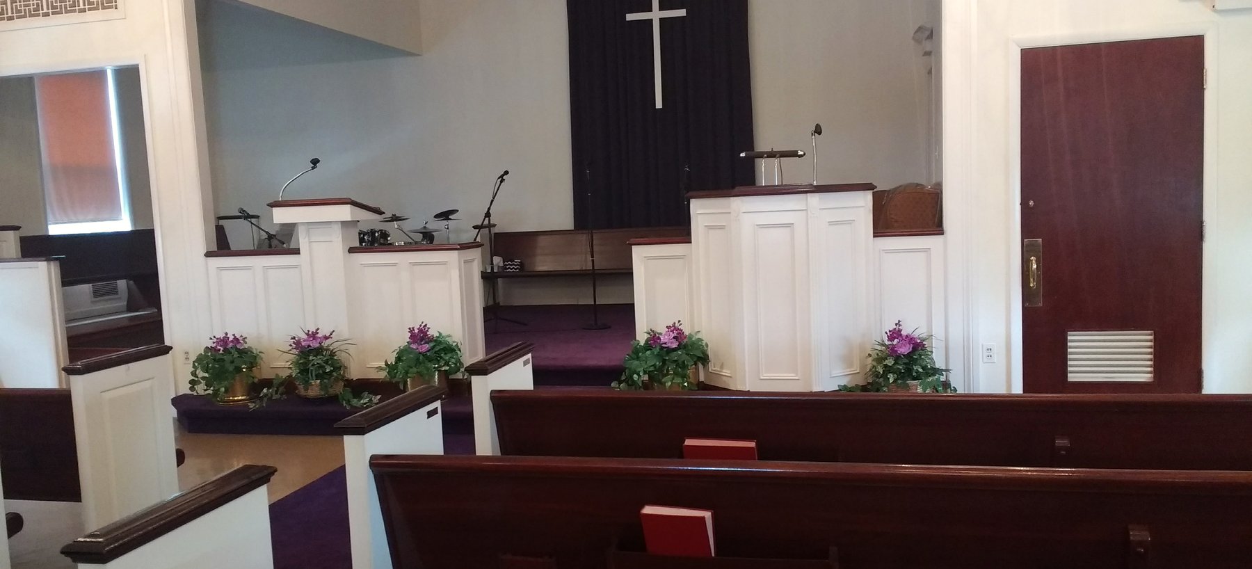 Inside the sanctuary: dark wood pews with purple cushions on a purple carpet, white panelled pulpit and lectern, pots of flowers along the front, and a white cross on a dark curtain behind.